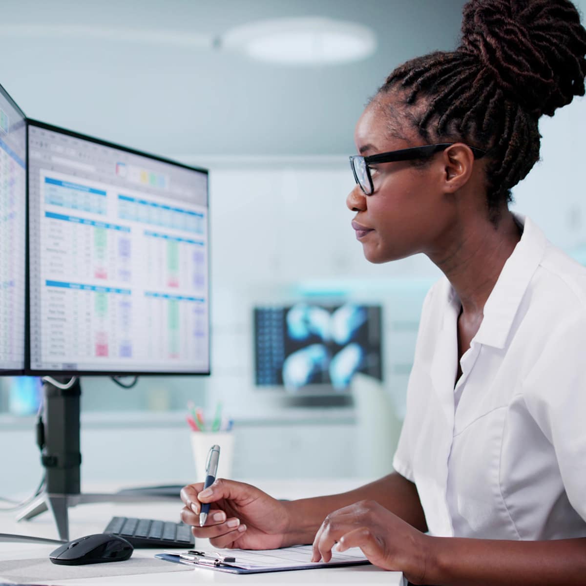 Healthcare professional reviewing computer monitors and writing notes at a workstation