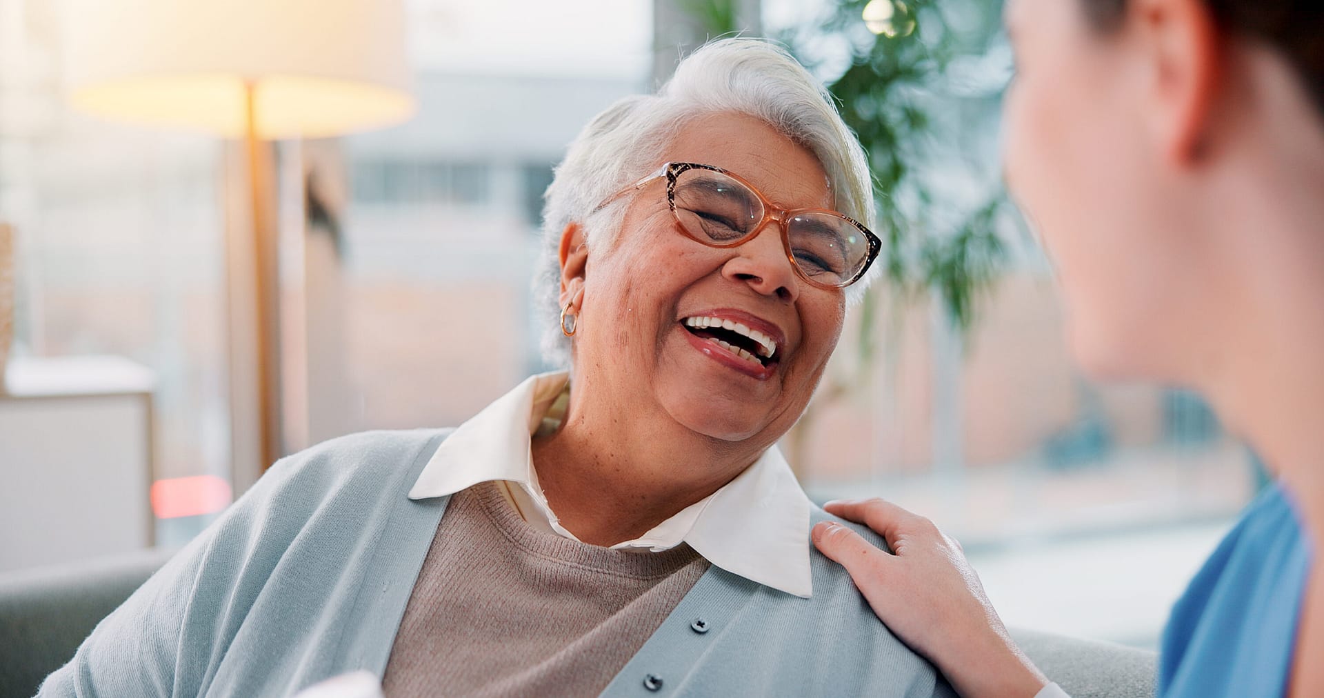 Older woman wearing glasses laughs as another person rests a hand on her shoulder
